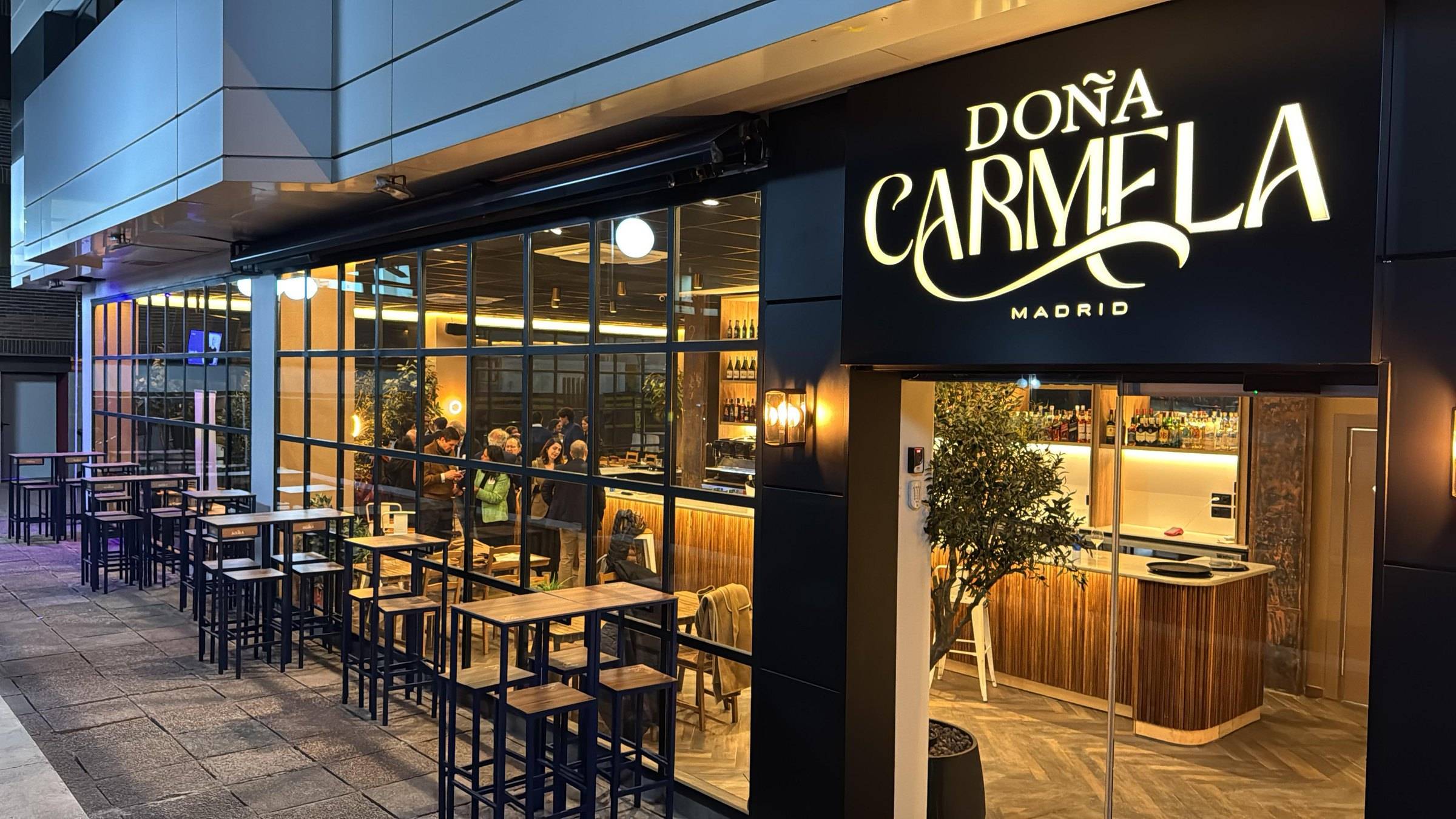 Fachada de Doña Carmela de noche en el Paseo de la Castellana, con el rótulo iluminado y la terraza de mesas altas