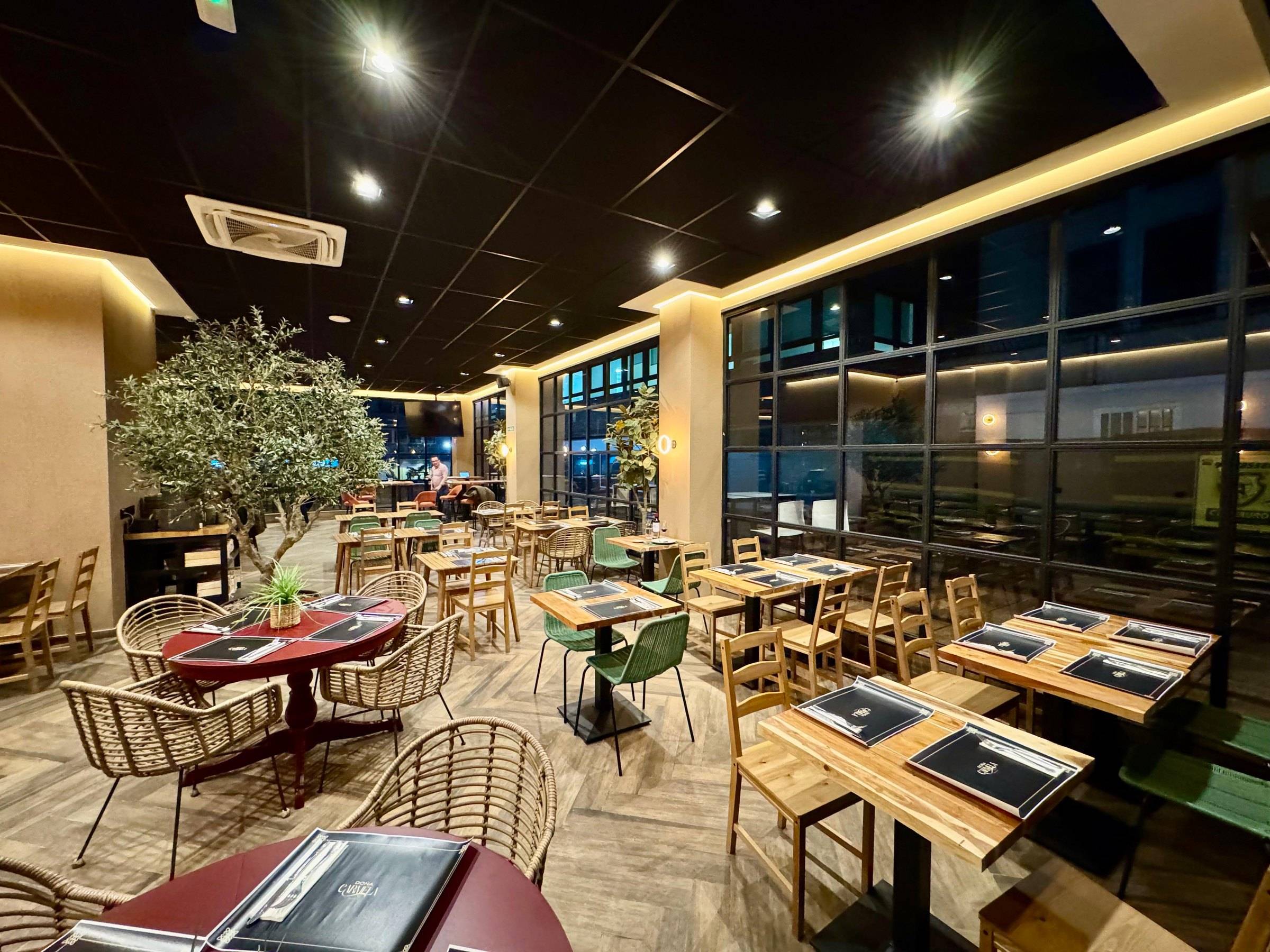 Salón de la planta alta de noche, con olivo, mesas de madera y grandes ventanales